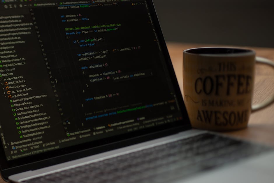 A close-up view of a laptop screen displaying lines of colorful code, positioned on a wooden desk. Next to the laptop, there is a ceramic mug with the words 'This coffee is making me awesome' printed