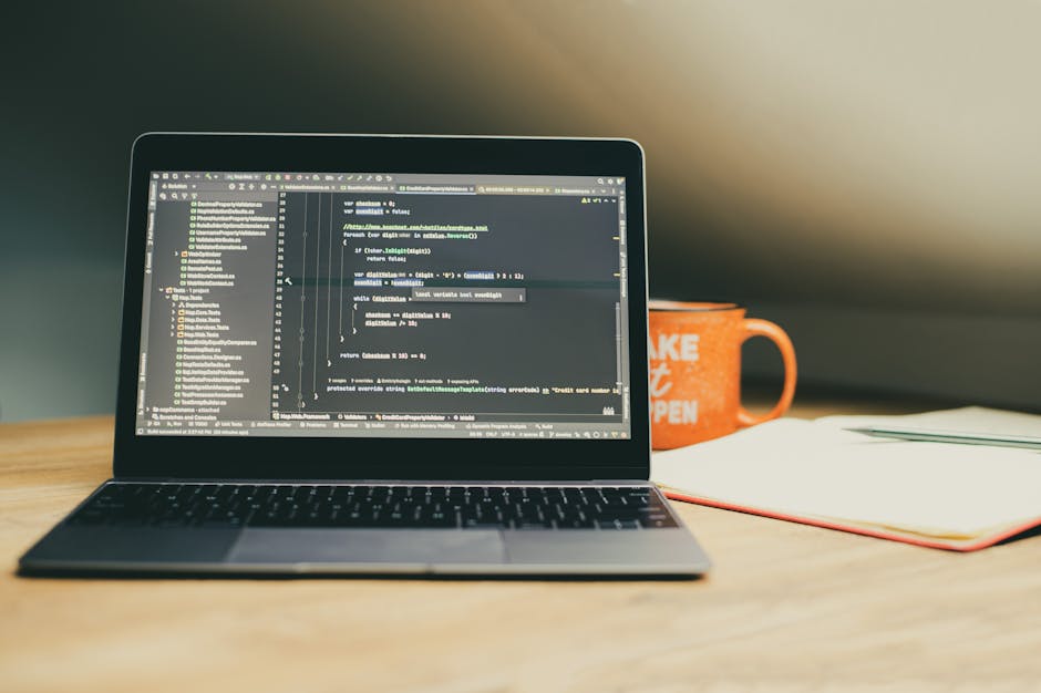 A close-up view of a laptop screen displaying lines of colorful code, positioned on a wooden desk. Next to the laptop, there is a ceramic mug with the words 'This coffee is making me awesome' printed