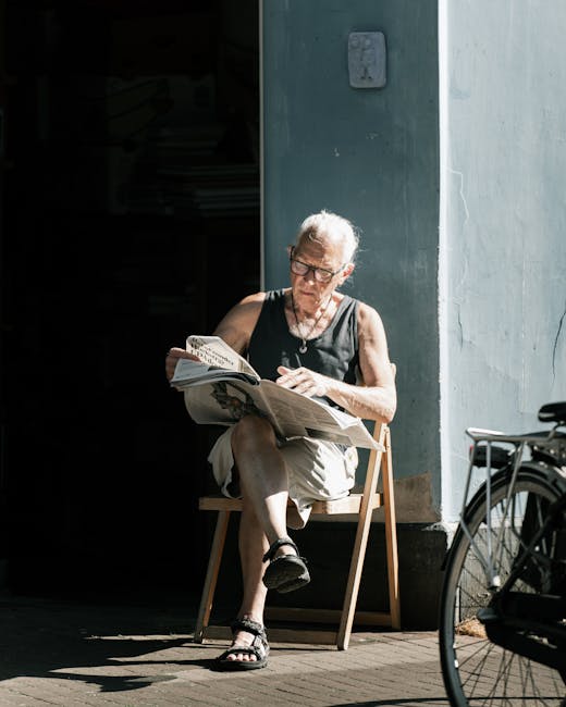 A senior woman with short, gray hair and glasses is sitting on a light wooden chair outside a building, reading a newspaper. She is dressed in a black sleeveless top, beige shorts, and black sandals. 